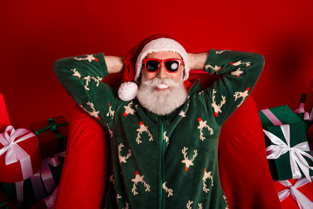 Santa in green reindeer Pajama relaxes with sunglasses among Christmas gifts in a red festive settingの写真素材