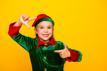 Young girl dressed as a cheerful Christmas elf pointing with a joyful expression on a bright yellow backgroundの写真素材