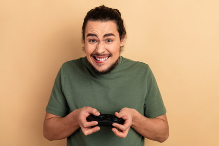Young man with green t shirt playing video game smiling while holding a controller against beige...