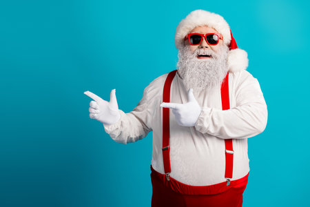 Santa Claus in red suspenders and white beard points to the right against a blue background wearing a hat and glassesの写真素材