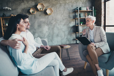 Photo of young couple hugging sit sofa psychotherapist office mature woman family counseling have conversation therapy sessionの写真素材