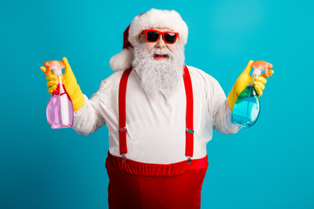 Santa in bright studio holding pink and blue spray bottles with yellow gloves and red suspenders for festive cleaning and holiday cheerの写真素材