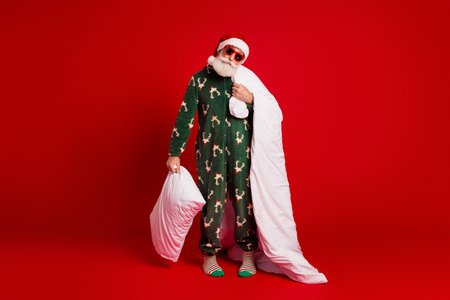 Playful Santa in festive pajama with blanket and pillow against a bold red backdropの写真素材