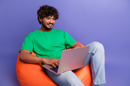 Young man in casual attire engaging with laptop while seated on an orange beanbag against a vibrant...