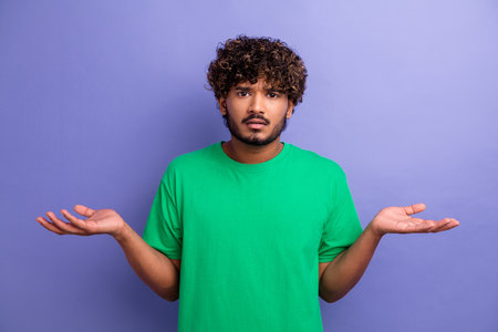Young man in casual green t-shirt gesturing in confusion with raised hands against a vivid purple backgroundの写真素材