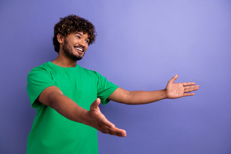 Confident young man in a green t-shirt gesturing with open arms in front of a violet-purple backgroundの写真素材