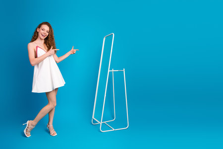 Young woman in a white dress stands by a minimal metal rack against a bright blue background smiling and pointing playfully at the scene creating a fun fashionable lifestyle momentの写真素材