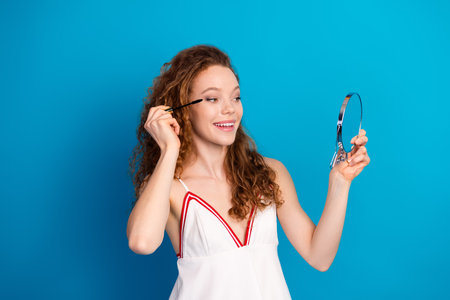 Young woman applying mascara and looking into a mirror wearing white dress with red trim against blue background for beauty fashionの写真素材