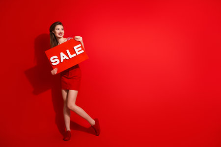 Energetic young woman in vibrant red attire displaying a sale sign on a bold red background for advertisementの写真素材