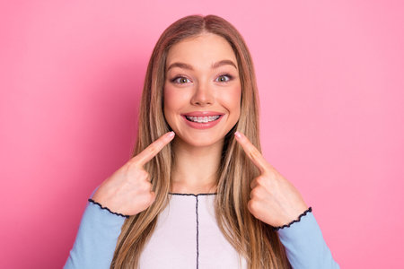 Happy young woman showcasing her dental braces with a cheerful smile in a pink background studio...
