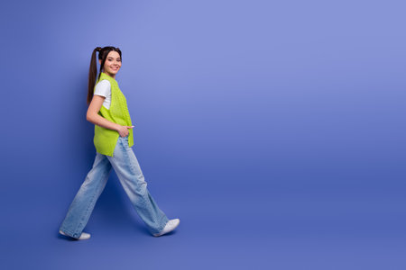 Young female student with lime vest walks along a blue studio backdrop smiling and showcasing casual fashion and lifestyleの写真素材
