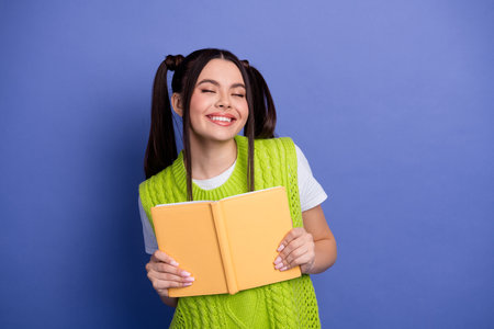 Cheerful young student girl in bright green vest smiles while reading an orange notebook against a...