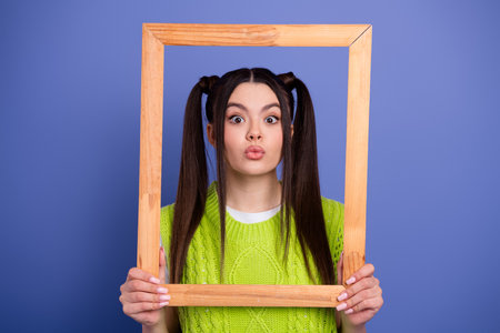 Young brunette girl in a bright green knit sweater frames her face with a wooden frame and playful...