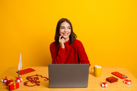 Funny christmas girl sitting at a bright yellow desk with laptop gifts beads and mug smiling at holiday magicの写真素材