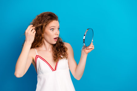 Young woman with curly brown hair checks her reflection in a round mirror against a bright blue background looking surprisedの写真素材