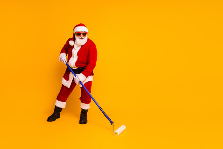 Santa with paint roller in bright yellow studio christmas scene playful grandpa santa claus costume fun creative holiday moodの写真素材