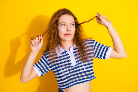 Playful young woman with curly red hair pulls her hair against a bright yellow background wearing a striped blue and white crop topの写真素材
