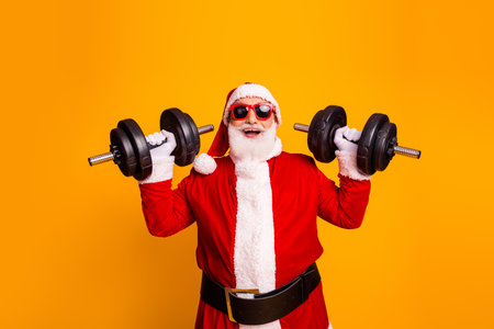 Santa lifts heavy dumbbells in red suit with white trim smiling against a bright orange background showcasing holiday strength and cheerの写真素材