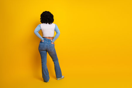 Rear view of a stylish young woman in a casual outfit standing against a vibrant yellow background...
