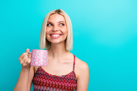 Happy young woman with blonde bob holding a pink mug smiles against teal blue background for lifestyle fashion and advertising useの写真素材