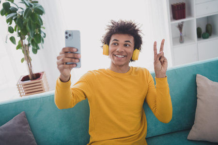 Photo of nice young man smart phone take selfie v-sign wear yellow pullover white interior apartment...