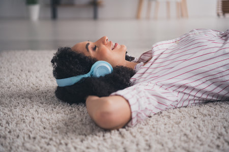 Photo of pretty cheerful lady wear casual shirt earphones arms behind head listening melodies indoors room home houseの写真素材