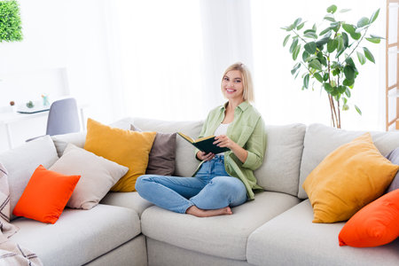 Photo of cheerful positive girl sitting on comfy sofa studying from home reading book homework...