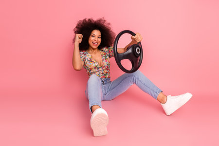 Joyful young woman holding a car steering wheel, seated on pink background and expressing excitement with a big smileの写真素材