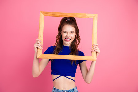 Cheerful young woman posing playfully with a wooden frame against a vibrant pink backdropの写真素材