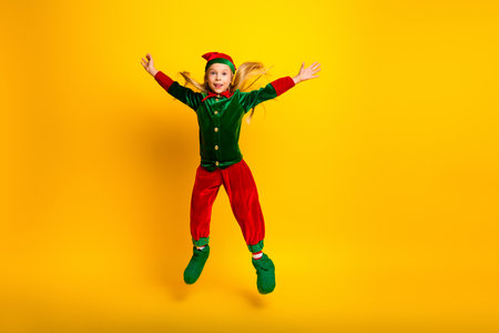 Energetic young child dressed as an elf jumping in festive excitement against a vibrant yellow...