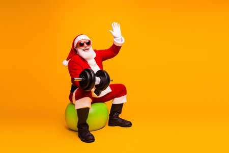 Santa lifts weights in a bright yellow studio wearing a red costume and hat with a dumbbell on a green ball a fun holiday fitness concept for Christmas and New Yearの写真素材