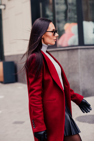 Stylish woman in red coat walking outdoors in urban city setting exuding confidence and fashionable demeanor during daylightの写真素材