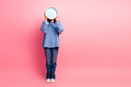 Young girl in a blue sweater holds a clock against a pink background conveying time fashion lifestyle and advertising potential for stock photographyの写真素材