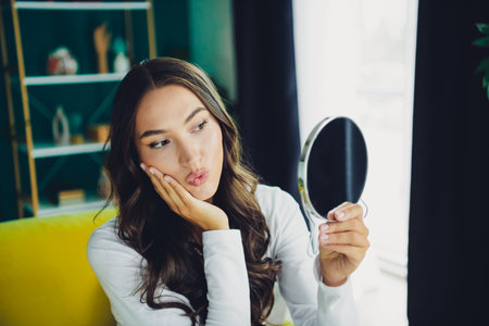 Woman sits on a couch and smiles softly while looking into a round mirror in a bright home interiorの写真素材