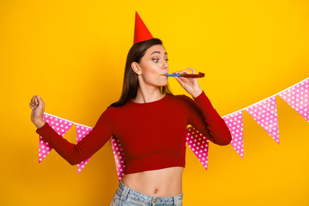 Young woman celebrates birthday party with red top party hat and pink banners on a bright yellow backgroundの写真素材