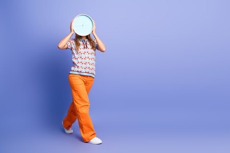 Young woman holds clock over her face in a playful fashion scene against a purple background conveying time pressure and cheerful style for advertising and lifestyleの写真素材