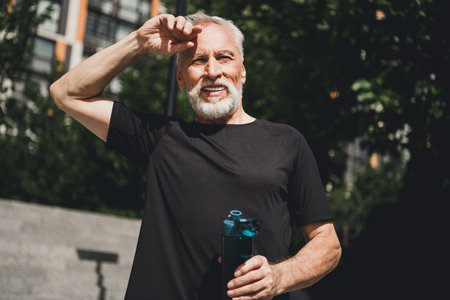Senior bearded man in black athletic shirt outdoors in a city park shielding his eyes while holding a water bottle during a morning workoutの写真素材
