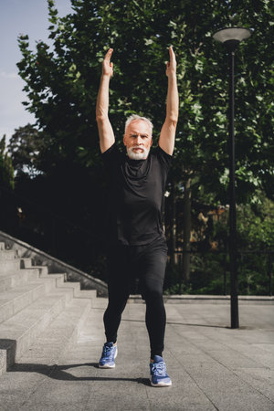 Active senior man with gray beard jumps outdoors in park during morning workout wearing modern sportswear for fitness and wellnessの写真素材