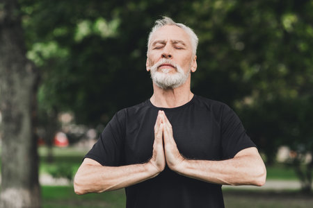 Senior bearded man meditates in a park outdoors with hands pressed in prayer and a peaceful expression as he practices mindfulness in a calm wellness momentの写真素材