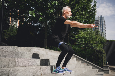 A senior bearded man in modern sportswear jumps on urban outdoor stairs during a morning park workout in the cityの写真素材