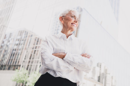 Confident elderly businesswoman stands in modern cityscape smiling with arms crossed as she proudly leads professional journey in the megapolisの写真素材