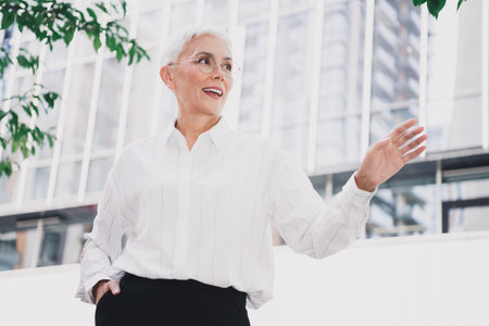 Senior businesswoman smiling outdoors in a modern city waving and engaging in professional style during daylightの写真素材