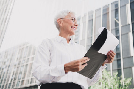 Confident elderly businesswoman outdoors in a modern city holding documents and smiling under daylight as she walks past glass buildings and urban architecture near a corporate skylineの写真素材
