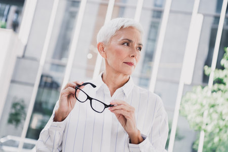 Confident elderly woman in a white striped shirt holds glasses in an urban cityscape ready for business and styleの写真素材