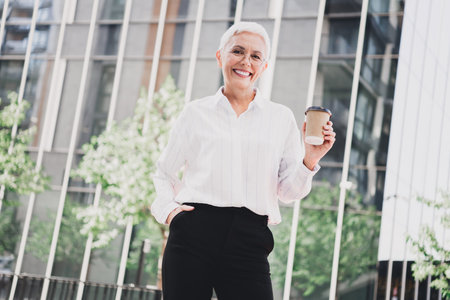 Confident elderly woman in white shirt and black pants stands outdoors in a modern city holding a coffee to go exuding professional eleganceの写真素材