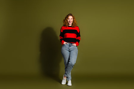 Young woman in red and black striped sweater jeans smiling against olive green backdrop for lifestyle fashion advertisingの写真素材