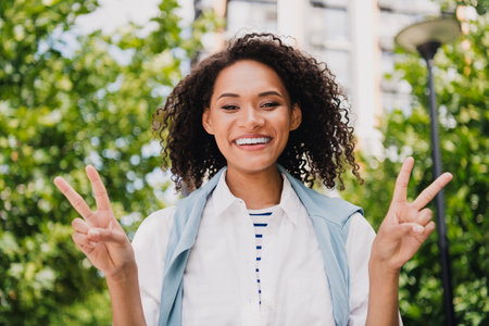 Smiling young woman outdoors in city park posing with peace signs wearing white shirt light jacket striped top for casual stylish lifestyle stock photoの写真素材