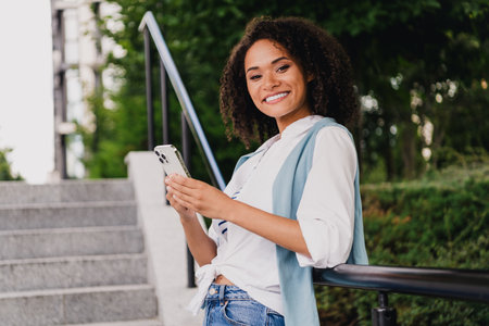 Smiling young woman with phone outdoors by steps in a modern urban park wearing casual fashion and relaxed lifestyle vibeの写真素材
