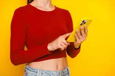 Young woman in red crop top uses yellow smartphone against bright yellow background showcasing casual fashion lifestyle and shopping promotionの写真素材