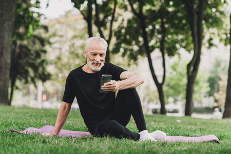 Older man with beard sits on a pink mat in the park outdoors using a smartphone during a morning workout in the city for wellness and fitness lifestyleの写真素材